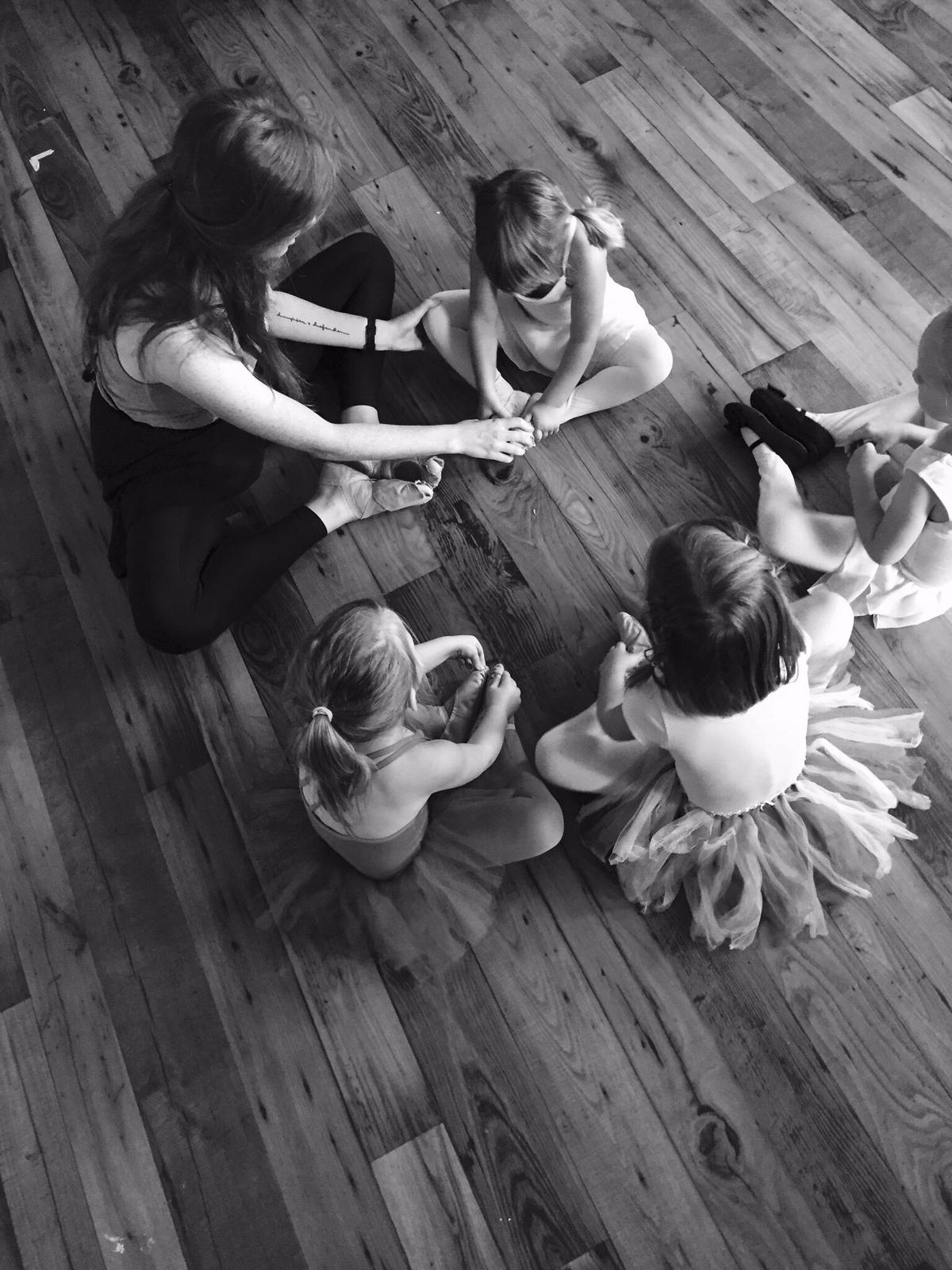 A teacher sitting on the studio floor with four young dancers, working through a stretch with them
