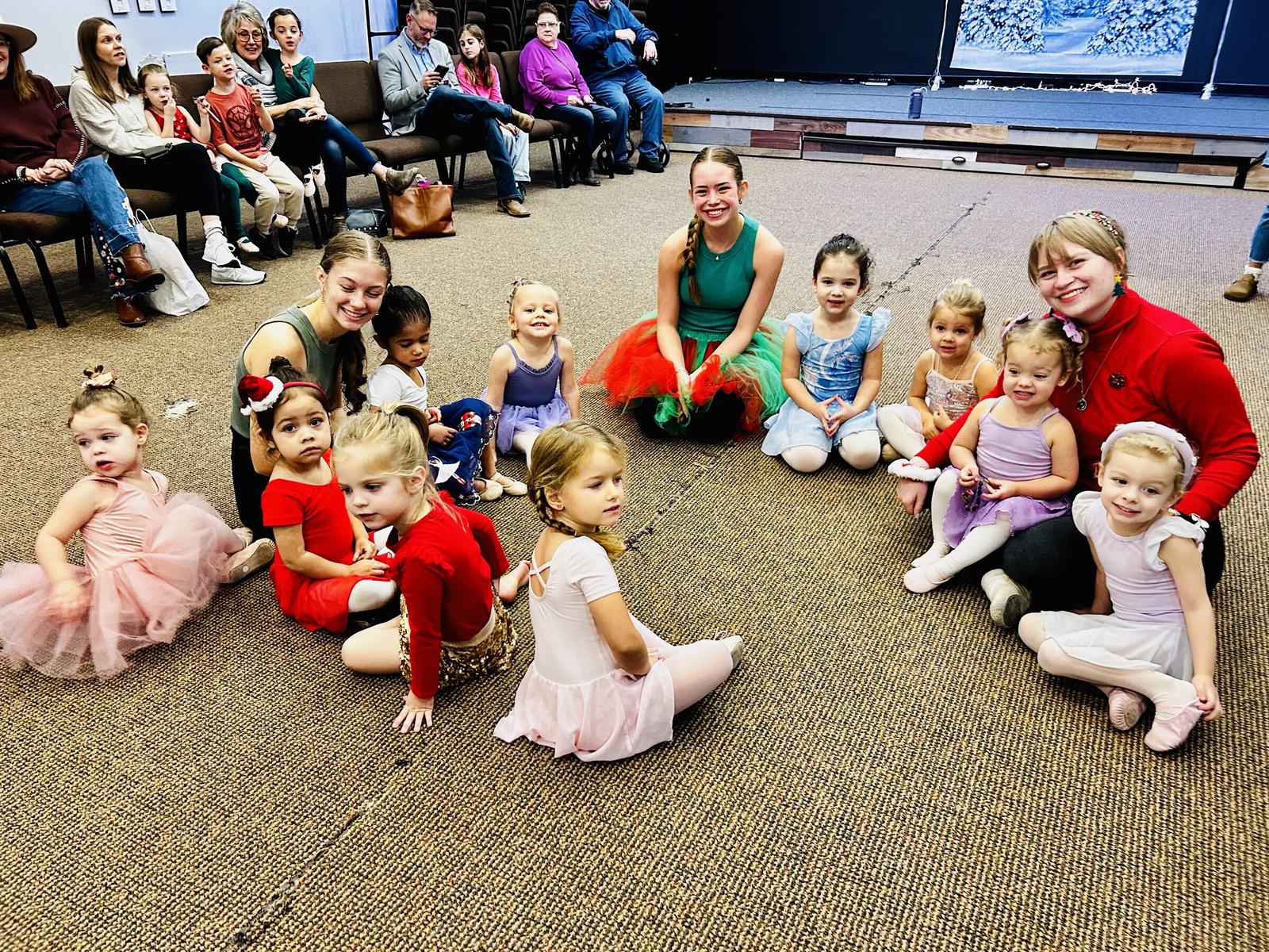 Dancers sitting on the floor at Watch Me Week, with families watching from chairs behind them